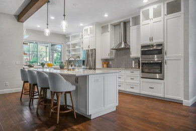 Mid-sized transitional u-shaped dark wood floor and brown floor open concept kitchen photo with a farmhouse sink, shaker cabinets, white cabinets, quartz countertops, gray backsplash, glass tile backsplash, stainless steel appliances, a peninsula and white countertops