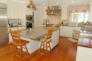 Example of a mid-sized u-shaped medium tone wood floor eat-in kitchen design in Bridgeport with a farmhouse sink, raised-panel cabinets, white cabinets, granite countertops, beige backsplash, stone tile backsplash, stainless steel appliances and an island