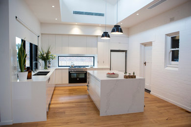 Große Moderne Wohnküche in L-Form mit weißen Schränken, Quarzwerkstein-Arbeitsplatte, Kücheninsel, flächenbündigen Schrankfronten, weißen Elektrogeräten, braunem Holzboden, Doppelwaschbecken und Rückwand-Fenster in Melbourne