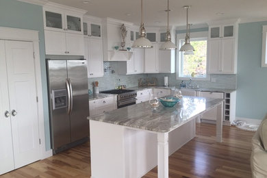 Example of a mid-sized beach style l-shaped light wood floor and brown floor open concept kitchen design in Wilmington with a farmhouse sink, shaker cabinets, white cabinets, marble countertops, white backsplash, stone tile backsplash, stainless steel appliances and an island