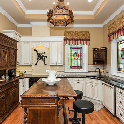 Example of a classic medium tone wood floor kitchen design in St Louis with a double-bowl sink, raised-panel cabinets, white cabinets, white backsplash, subway tile backsplash, stainless steel appliances, an island and black countertops