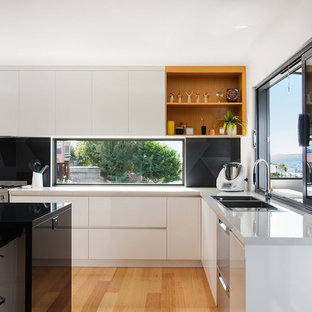 Ejemplo de cocina comedor de galera, minimalista, de tamaño medio, con fregadero encastrado, puertas de armario blancas, encimera de laminado, salpicadero de vidrio, electrodomésticos de acero inoxidable, suelo laminado y una isla