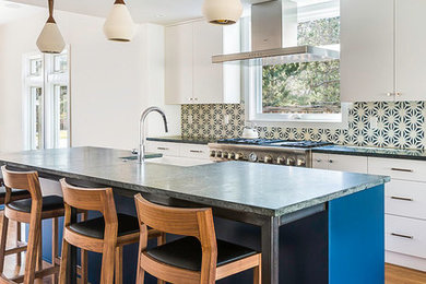 Example of a trendy medium tone wood floor and brown floor eat-in kitchen design in Denver with a farmhouse sink, flat-panel cabinets, white cabinets, stainless steel appliances, an island, soapstone countertops, blue backsplash, cement tile backsplash and gray countertops