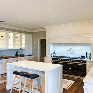 Design ideas for a transitional u-shaped eat-in kitchen in Melbourne with a farmhouse sink, recessed-panel cabinets, white cabinets, white splashback, subway tile splashback, black appliances, dark hardwood floors, with island, brown floor and white benchtop.
