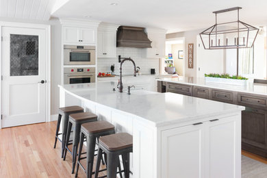Example of a farmhouse u-shaped light wood floor and beige floor kitchen design in Boston with a farmhouse sink, shaker cabinets, white cabinets, white backsplash, subway tile backsplash, stainless steel appliances, two islands and white countertops