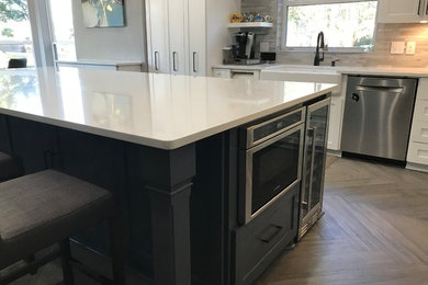 Example of a large transitional medium tone wood floor and brown floor open concept kitchen design in Houston with a farmhouse sink, shaker cabinets, white cabinets, quartz countertops, gray backsplash, porcelain backsplash, stainless steel appliances, an island and white countertops