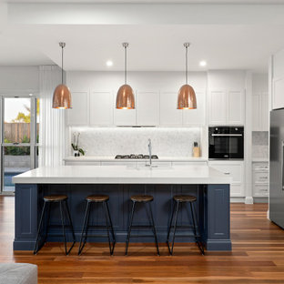 Inspiration for a mid-sized beach style single-wall kitchen in Brisbane with a farmhouse sink, shaker cabinets, white cabinets, quartz benchtops, grey splashback, marble splashback, stainless steel appliances, medium hardwood floors, with island, multi-coloured floor and white benchtop.