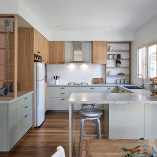 This is an example of a contemporary u-shaped eat-in kitchen in Melbourne with a double-bowl sink, flat-panel cabinets, grey cabinets, white splashback, subway tile splashback, white appliances, medium hardwood floors, with island, brown floor and multi-coloured benchtop.