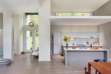 Inspiration for a medium sized modern l-shaped kitchen in Bridgeport with a submerged sink, flat-panel cabinets, grey cabinets, engineered stone countertops, white splashback, glass tiled splashback, stainless steel appliances, light hardwood flooring, an island, grey floors and white worktops.