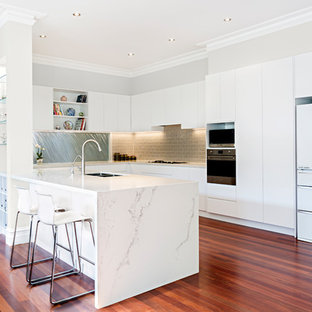 Design ideas for a contemporary u-shaped kitchen in Melbourne with a double-bowl sink, flat-panel cabinets, white cabinets, brown splashback, subway tile splashback, white appliances, medium hardwood floors, a peninsula and red floor.
