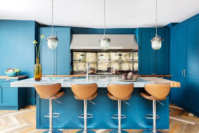 Photo of a classic l-shaped kitchen/diner in London with a belfast sink, shaker cabinets, blue cabinets, metallic splashback, mirror splashback, integrated appliances, medium hardwood flooring, an island, beige floors and grey worktops.