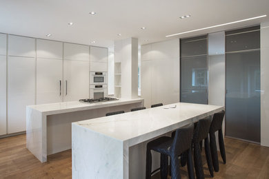 Large trendy l-shaped medium tone wood floor and brown floor eat-in kitchen photo in Montreal with white cabinets, marble countertops, white appliances, two islands and flat-panel cabinets