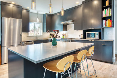 Example of a trendy l-shaped light wood floor kitchen design in Seattle with flat-panel cabinets, white backsplash, stainless steel appliances, an island and gray countertops