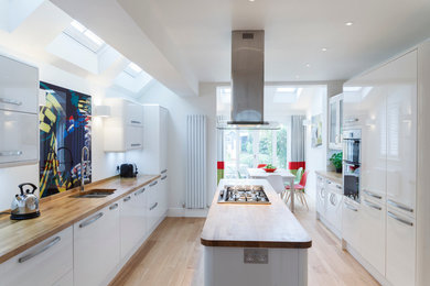 Design ideas for a contemporary galley kitchen/diner in Cambridgeshire with a submerged sink, flat-panel cabinets, white cabinets, wood worktops, stainless steel appliances, light hardwood flooring, an island and multi-coloured splashback.