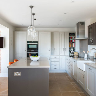 Medium sized contemporary grey and white l-shaped open plan kitchen in Buckinghamshire with shaker cabinets, grey cabinets, an island, a belfast sink, grey splashback, black appliances, beige floors and grey worktops.