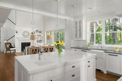 Mid-sized trendy l-shaped medium tone wood floor and brown floor open concept kitchen photo in Los Angeles with a farmhouse sink, recessed-panel cabinets, white cabinets, marble countertops, white backsplash, subway tile backsplash, white appliances, an island and white countertops