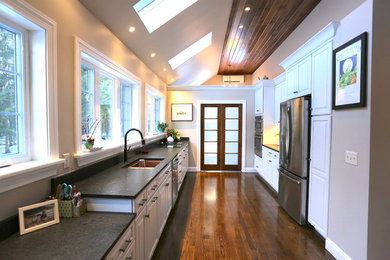 Huge trendy galley medium tone wood floor and brown floor eat-in kitchen photo in Portland Maine with an undermount sink, raised-panel cabinets, white cabinets, granite countertops, black backsplash, stone slab backsplash, stainless steel appliances, an island and black countertops