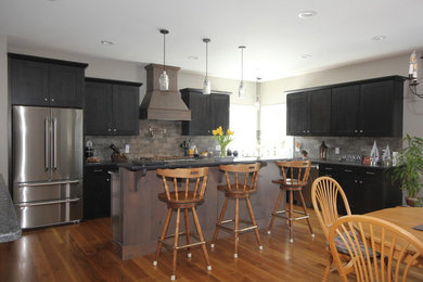 Mid-sized cottage l-shaped medium tone wood floor and brown floor open concept kitchen photo in Boston with an island, granite countertops, gray backsplash, stone tile backsplash, stainless steel appliances, black countertops, a farmhouse sink, shaker cabinets and dark wood cabinets