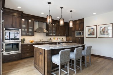 Mid-sized arts and crafts l-shaped light wood floor and brown floor open concept kitchen photo in Minneapolis with a farmhouse sink, shaker cabinets, dark wood cabinets, quartz countertops, beige backsplash, stainless steel appliances, an island and subway tile backsplash