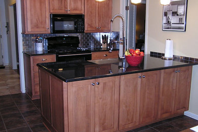 Klassisk inredning av ett litet kök, med en rustik diskho, skåp i shakerstil, skåp i mellenmörkt trä, granitbänkskiva, stänkskydd med metallisk yta, stänkskydd i stenkakel, svarta vitvaror, klinkergolv i porslin, en halv köksö och brunt golv