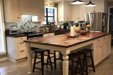 Photo of a large rural l-shaped enclosed kitchen in New Orleans with a belfast sink, beaded cabinets, white cabinets, grey splashback, stone tiled splashback, stainless steel appliances, an island, beige floors and black worktops.