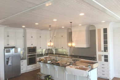 Large beach style l-shaped medium tone wood floor eat-in kitchen photo in Miami with a farmhouse sink, shaker cabinets, white cabinets, granite countertops, green backsplash, stainless steel appliances and an island