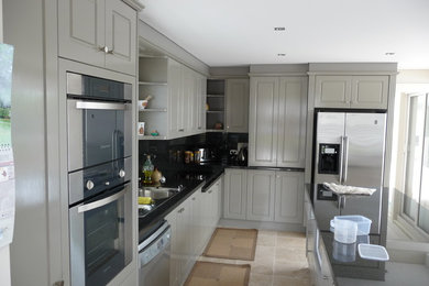 Cette image montre une cuisine design en L avec un placard à porte affleurante, des portes de placard grises, une crédence noire, un électroménager en acier inoxydable, un sol en travertin et îlot.
