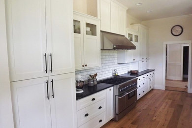 Example of a mid-sized classic galley medium tone wood floor eat-in kitchen design in Seattle with a farmhouse sink, shaker cabinets, white cabinets, soapstone countertops, white backsplash, ceramic backsplash, stainless steel appliances and no island