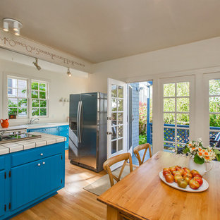 This is an example of an arts and crafts galley eat-in kitchen in San Francisco with louvered cabinets, blue cabinets, tile benchtops, stainless steel appliances, light hardwood floors and with island.