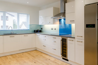 This is an example of a medium sized contemporary l-shaped kitchen/diner in Other with a built-in sink, flat-panel cabinets, white cabinets, granite worktops, blue splashback, glass sheet splashback, integrated appliances and light hardwood flooring.
