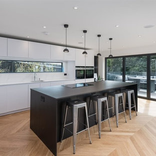 Design ideas for a medium sized contemporary l-shaped kitchen in Sussex with a single-bowl sink, flat-panel cabinets, white cabinets, window splashback, black appliances, light hardwood flooring, an island and white worktops.