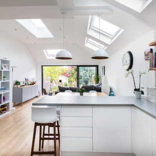 Photo of a contemporary l-shaped open plan kitchen in Essex with flat-panel cabinets, white cabinets, white splashback, light hardwood flooring, a breakfast bar, beige floors and grey worktops.