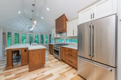 Large classic galley kitchen/diner in Indianapolis with a belfast sink, raised-panel cabinets, medium wood cabinets, engineered stone countertops, white splashback, glass tiled splashback, stainless steel appliances, light hardwood flooring, an island, brown floors, white worktops and a vaulted ceiling.