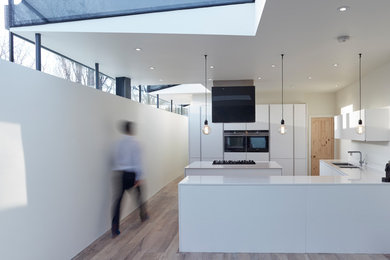 Example of a trendy l-shaped light wood floor and beige floor kitchen design in London with a double-bowl sink, flat-panel cabinets, white cabinets, white backsplash, black appliances, a peninsula and white countertops