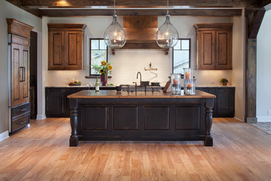 This is an example of a medium sized traditional l-shaped open plan kitchen in Miami with a belfast sink, raised-panel cabinets, dark wood cabinets, wood worktops, white splashback, ceramic splashback, integrated appliances, light hardwood flooring, an island and brown floors.