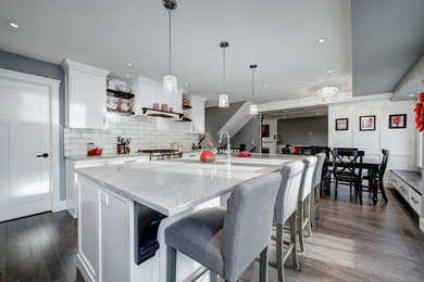 Large classic l-shaped kitchen/diner in Calgary with a belfast sink, shaker cabinets, white cabinets, engineered stone countertops, white splashback, metro tiled splashback, stainless steel appliances, medium hardwood flooring, an island and brown floors.