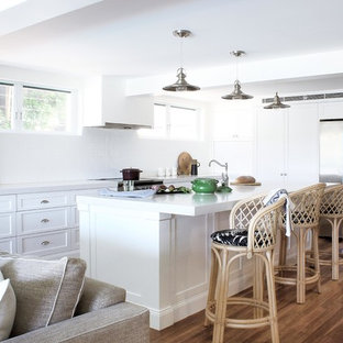This is an example of a beach style l-shaped open plan kitchen in Sydney with a farmhouse sink, shaker cabinets, white cabinets, granite benchtops, white splashback, ceramic splashback, stainless steel appliances, with island, white benchtop, medium hardwood floors and brown floor.