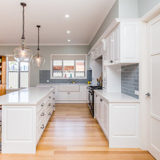 Photo of a mid-sized country l-shaped kitchen in Geelong with an integrated sink, raised-panel cabinets, white cabinets, grey splashback, ceramic splashback, medium hardwood floors, multiple islands and brown floor.