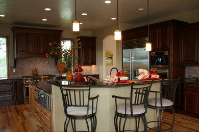 Large mountain style medium tone wood floor kitchen photo in Denver with raised-panel cabinets, dark wood cabinets, granite countertops, glass tile backsplash, stainless steel appliances and an island