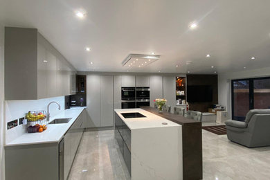 Inspiration for an expansive contemporary l-shaped open plan kitchen in London with a submerged sink, flat-panel cabinets, grey cabinets, quartz worktops, white splashback, stone slab splashback, black appliances, porcelain flooring, a breakfast bar, grey floors and white worktops.