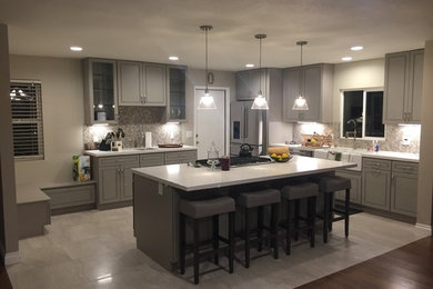 Photo of a large classic l-shaped kitchen in Los Angeles with a belfast sink, raised-panel cabinets, grey cabinets, composite countertops, grey splashback, stone tiled splashback, stainless steel appliances, an island, grey floors and white worktops.