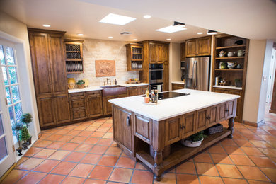 Mid-sized mountain style l-shaped terra-cotta tile and brown floor eat-in kitchen photo in Phoenix with a farmhouse sink, raised-panel cabinets, medium tone wood cabinets, marble countertops, white backsplash, stone tile backsplash, stainless steel appliances and an island