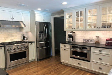 Enclosed kitchen - mid-sized transitional l-shaped light wood floor and beige floor enclosed kitchen idea in Boston with a farmhouse sink, recessed-panel cabinets, white cabinets, granite countertops, white backsplash, marble backsplash, stainless steel appliances, no island and black countertops