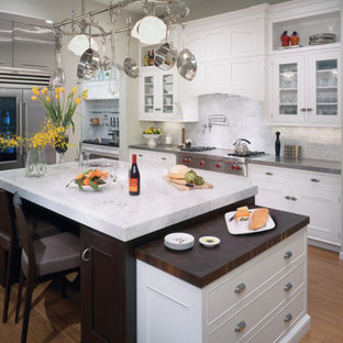 Design ideas for a large traditional l-shaped eat-in kitchen in Seattle with a triple-bowl sink, glass-front cabinets, white cabinets, white splashback, ceramic splashback, stainless steel appliances, light hardwood floors, with island, marble benchtops and brown floor.