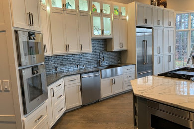 Example of a mid-sized transitional l-shaped medium tone wood floor and brown floor open concept kitchen design in Other with a farmhouse sink, shaker cabinets, white cabinets, quartzite countertops, gray backsplash, glass tile backsplash, stainless steel appliances, an island and white countertops