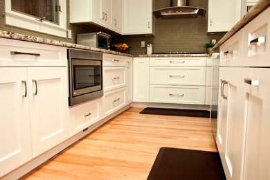 Mittelgroße Moderne Wohnküche in L-Form mit Schrankfronten mit vertiefter Füllung, weißen Schränken, Granit-Arbeitsplatte, bunter Rückwand, Rückwand aus Glasfliesen, Küchengeräten aus Edelstahl, hellem Holzboden und Kücheninsel in Detroit