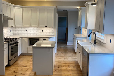 Example of a mid-sized farmhouse l-shaped light wood floor and white floor kitchen pantry design in Other with a farmhouse sink, shaker cabinets, white cabinets, quartz countertops, white backsplash, subway tile backsplash, stainless steel appliances, an island and gray countertops