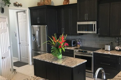 This is an example of a medium sized traditional u-shaped enclosed kitchen in Orlando with black cabinets, grey splashback, an island, a belfast sink, shaker cabinets, granite worktops, glass tiled splashback, stainless steel appliances, travertine flooring and beige floors.
