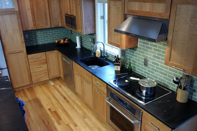 Example of a mid-sized transitional l-shaped light wood floor and brown floor kitchen design in Cedar Rapids with an undermount sink, shaker cabinets, light wood cabinets, soapstone countertops, green backsplash, subway tile backsplash, stainless steel appliances and an island