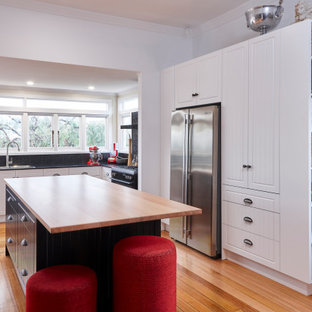 Design ideas for a country l-shaped kitchen in Other with raised-panel cabinets, white cabinets, black splashback, stainless steel appliances, medium hardwood floors, with island, brown floor and black benchtop.